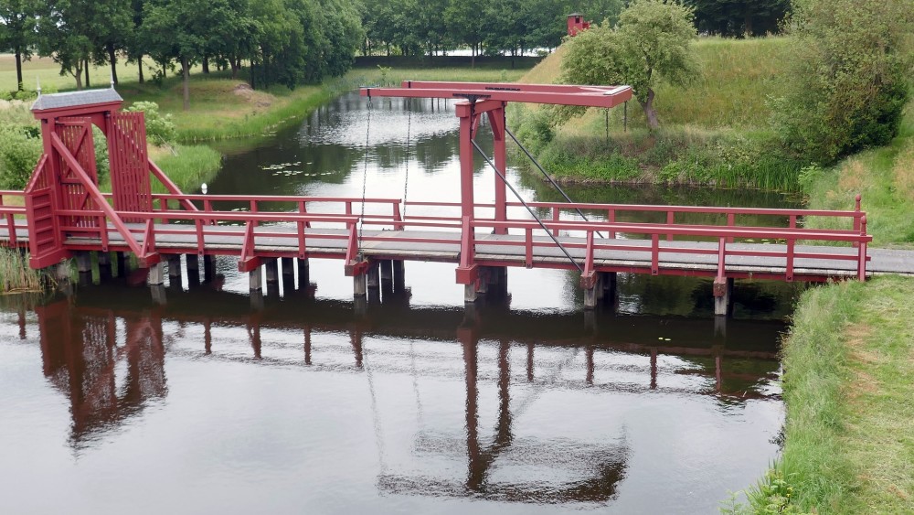 Een rode brug, waar je overheen kan lopen tijdens jouw vakantie in Groningen, die het vaste land verbind met de vestingstad Bourtange. 